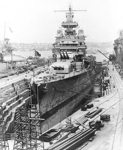 A raised warship in a drained dock surrounded by construction equipment