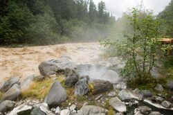 Steaming pool of water surrounded by a group of rocks next to a muddy river.