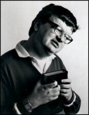 A black and white photo of a white man with an unusually large head, expressionless face and short hair looking to the right. He wears glasses and a white collared shirt with a dark sweater over it. He holds a closed book in both hands.