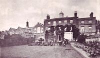 A sepia photograph of the back of a Georgian house and schoolrooms. Edwardian schoolboys and their masters can be seen on the grass in front.