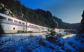 A train of two-level passenger rail cars at a station. The coaches are stainless steel, with red and blue horizontal stripes halfway up their sides.