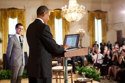 Obama in blue suit standing at a podium in front of an audience as man in a light grey suit looks on.