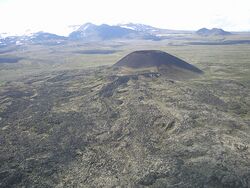 A barren plateau with a small cone-shaped mound in the foreground, a low-lying glaciated mountain in the left background and a small elongated mound in the right background.
