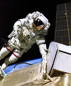 An astronaut in a white spacesuit with a red, white, and blue flag on the left shoulder in the foreground with a black background