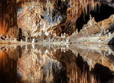 Mirror-like water in a cave. Across from the camera, at the shore, rests a number of stalactites and stalagmites