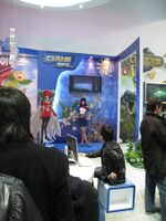 Inside a large, brightly lit convention center room with white walls is positioned a promotional display booth for a video game. A saleswoman clad in a blue shirt and skirt and a red bowtie motions towards several illustrations on the booth, explaining their implications. The illustrations are anime-styled and depict several outlandish and brightly colored creatures. Three men in dark jackets watch the demonstration.