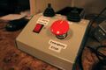 A closeup of a handmade electronic control box on a control room desk. The box has a slanted front–top face equipped with a red rocker switch labeled "3 degree Override" and a large red button labeled "Emergency STOP". The horizontal top face of the box has two identical small black joysticks with rubber bellows, labeled with directions. Some of the labels are hidden and/or blurry, but the left joystick appears to be labeled "UP/DOWN/LEFT/RIGHT" and the right joystick appears to be labeled "S/N/E/W" (compass directions rotated 180° from the usual arrangement). The box's sides are blue and its face is gray. The labels were printed by a label maker, and have black text on a white background.