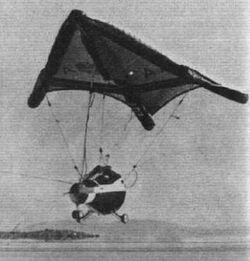 A spacecraft on wheels, suspended from a large triangular wing, in flight close to the ground.