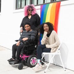 Pictured are three Black and disabled people. On the left is a non-binary person holding a cane, in the middle there is a woman sitting in a power wheelchair, and on the right is a woman sitting in a chair. They are all partially smiling at the camera while a rainbow pride flag drapes on the wall behind them.