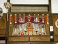 A large, decorative alcove shrine, with red paper lanterns decorated with black text strung across the top, a miniature torii gate in front and two red daruma dolls at either side.