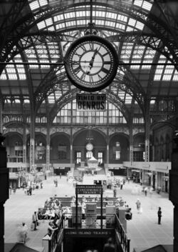Large high-ceilinged space with round iron arches, covered in glass-dome skylights, floored with vault lights, clock central in foreground.