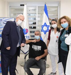 Reuven Rivlin and the first person to receive the BriLife vaccine look at the viewer alongside two nurses. The flag of Israel sits in the background.