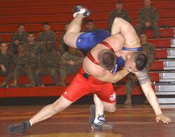 Two men wrestling in a gymnasium, watched by a group of uniformed soldiers
