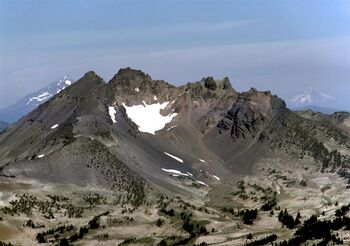 A highly eroded volcano with jagged cliffs, with two steep volcanoes in the background to its left and to its right.