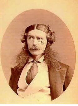 Head and shoulders portrait of a foppish Victorian gentleman wearing a monocle, with longish hair parted in the middle and curly at each side, exaggerated long bushy sideburns and a handlebar moustache. He wears a jacket with very wide lapels, an upright collar and wide tie tucked into his double breasted waistcoat.