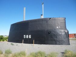 A very large submarine conning tower in a gravel park.
