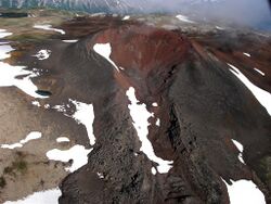 A dark-coloured volcanic cone rising above a sparsely snow-covered rocky plateau.