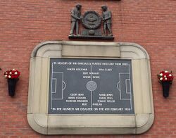 A stone tablet, inscribed with the image of a football pitch and several names. It is surrounded by a stone border in the shape of a football stadium. Above the tablet is a wooden carving of two men holding a large wreath.