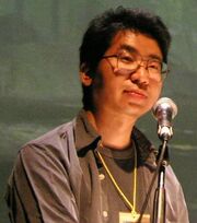 A bespectacled Japanese man wearing a black T-shirt and a gray jacket looks ahead with anticipation.