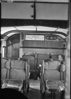 Photograph of the passenger cabin of a Short Kent airliner, showing pullman-style seating and a man sitting in the rearmost row, a map of the Genoa-Alexandria route on the wall.