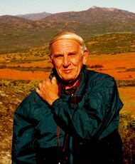 Edward M. Burgess pictured in Namaqualand, South Africa. Burgess is wearing a dark blue-green jacket and is carrying a camera strapped around his neck (tourist style). His left arm is positioned across his chest with his left hand on his right shoulder. Burgess is looking directly into the (active) camera with a slight smile. In the background are numerous mountainous peaks breaking to a pale blue sky at the top of the photo. At approximately shoulder height the green vegetation in the foot hills gives way to an expanse of orange-red flowers.