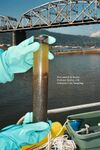 A diver’s blue-gloved hands holds a cylindrical sediment core. The core is inside a plastic tube through which dark sediments and murky water can be seen. Image text reads “McCormick & Baxter Portland Harbor, OR Sediment Core Sampling”. In the background there is the deck of a boat, water, a bridge, and a forested ridge.