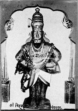 A black and white photograph of a stone icon of an arms-akimbo man standing on a brick and wearing a dhoti, angarkha (shirt), uparna (cloth that flows over both his hands), a crown, fish-shaped earrings and long necklaces till his waist.