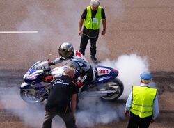 A silver and blue sport motorcycle with racing numbers and covered sponsor logos. The rider has a reflective helmet and wears brightly colored leathers also covered with logos. His hands on the clutch and brake levers while the front wheel does not turn and the rear wheel spins and emits a cloud of white smoke. Three officials stand close by and observe.