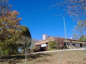 The visitor center is depicted as a building on a hill, surrounded by trees with autumn foliage and a clear blue sky.