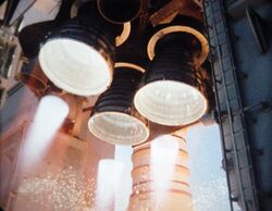 Three bell-shaped rocket engine nozzles projecting from the aft structure of a Space Shuttle orbiter. The cluster is arranged triangularly, with one engine at the top and two below, with two smaller nozzles visible to the left and right of the top engine. The three larger engines are firing, with white-hot flames visible projecting from each nozzle. The Space Shuttle's left solid rocket booster (a white, cylindrical rocket) is visible in the background, with the two large, grey tail service masts visible to the left and right of the orbiter's aft structure.