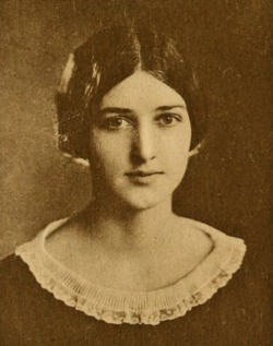 A young white woman with dark hair parted center, wearing a dark dress or blouse with a round lace-trimmed neckline