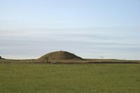 A small round hill lies beyond a field of tall grasses. The hill is also grass covered. A small entrance way to a passage into the hill is just visible at centre and a small human figure stands on top of the elevation.