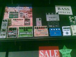 A music store display of effect pedals for bass is shown. The pedals have foot-operated switches to turn the effect on an off and knobs for controlling the tone.