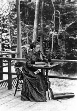 A white woman seated at a table outdoors looking into a microscope. She is wearing a dark dress and there is a forest in the background.