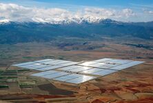 Concentrated solar power parabolic troughs in the distance arranged in rectangles shining on a flat plain with snowy mountains in the background