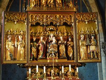 The central section of an elaborately carved, painted and gilded altarpiece showing the Virgin Mary and Christ Child seated in majesty and surrounded by saints and angels. Although the flesh and some details are painted in colour, most of the surfaces are gilt. The figures are all chubby-faced and have a charming quality.