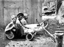 Trio of men seated on the ground with ankles shackled to a pole lying horizontally