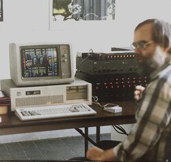 Photograph of bearded man in a plaid shirt sitting at desktop computer next to a sound mixing device.