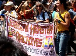 Image of three people walking in a crowd, smiling and holding a banner that reads "LESBIAN HERSTORY ARCHIVES".