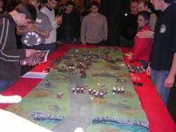 Several men surround a table, on which lies a small scale model of an undulating ground. Many small figures of monsters and medieval warriors and siege weapons are placed on that terrain. Dice are scattered among the figures.