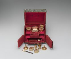A small, elaborate box, featuring a hinged lid, two swing doors at the front and a small pull-out drawer; the interior is entirely red and features small items that seem to be part of a toilette set