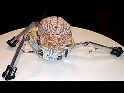 Photograph of model of a brain mounted on a three-legged robotic base with actuating mechanisms and exposed wires. The brain has a face with mouth and red eyes, and a small arm with grasping hands emerging from each side.