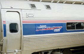 A stainless steel passenger rail car with a thick blue stripe across the windows and two thinner red stripes above. A logo within the blue stripe reads "Amtrak NortheastDirect Service".