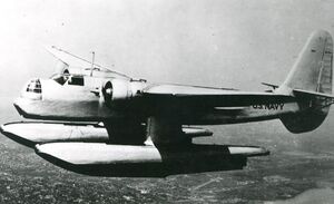 A twin-engined, monoplane aircraft, fitted with two large floats and with a large tail fin, flies over a city.