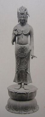 Frontal view of a statue on a pedestal with the left hand raised to breast height. The left hand carries a small object shaped like a vase or pot.