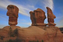 Four sandstone hoodoos with three of the four being closely-grouped and the fourth separated by a short distance, all standing atop a larger sandstone base, and lit by the setting sun.