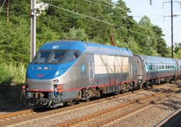 Stainless steel electric locomotive with a blue roof and a thin red sill stripe. Behind it are stainless steel passenger cars with a mottled blue window stripe and a thin red sill stripe.