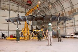 Yellow crane hoisting a wing of an aircraft, with several people standing below securing the wing. This takes place inside an aircraft hangar.