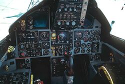 Cockpit of jet fighter with circular dials and gauges: A control stick protrude from between where the pilot's legs would be.