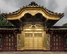 The front of a Shinto Shrine, colored gold and brown, with a golden overhang to the front doors. There are carvings both above and to the sides of the main doors. The sky is overcast and dark.